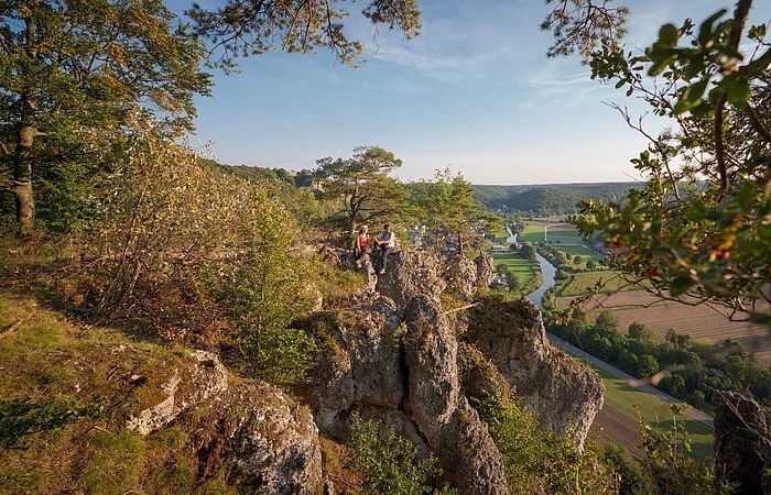 Ein Wanderpärchen, etwas entfernt, steht links auf einem Felsen an der Arnsberger Leite, die gut zu sehen ist. Dahinter erstreckt sich der Blick ins Tal. Der Himmel erstrahlt wolkenlos.