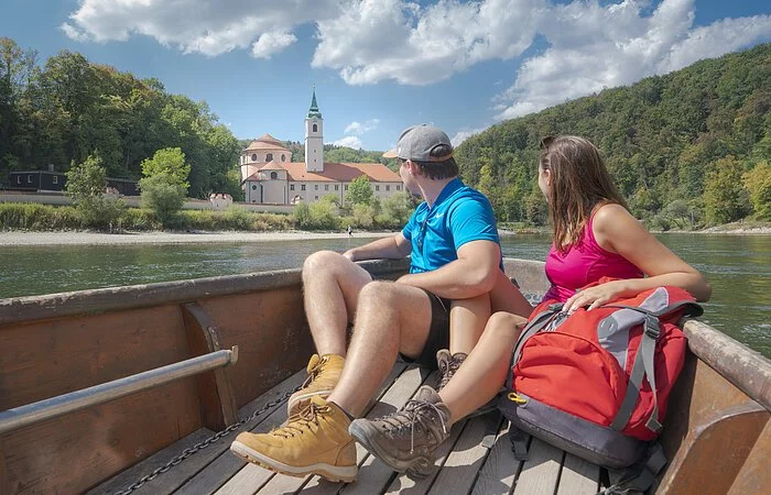 In dem hölzernen Boot sitzen ein Mann und eine Frau, beide in Wanderkleidung mit dem Blick Richtung Kloster Weltenburg gerichtet. Das Kloster ist im Hintergrund von weitem zu sehen umrahmt von grünen Wäldern.