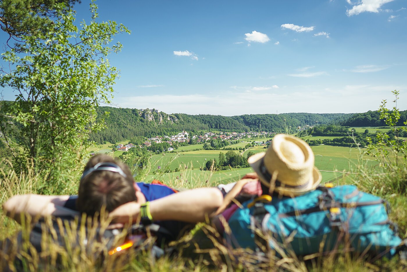 Ein Mann mit verschränkten Armen und daneben eine Frau mit Strohhut, die ihren Kopf auf ihren blauen Rucksack abgelegt hat, liegen im Gras. Sie genießen den Blick von der Arnsberger Leite ins Tal. Der Himmel strahlt blau und nur ein paar Wolken haben sich dazu verirrt.