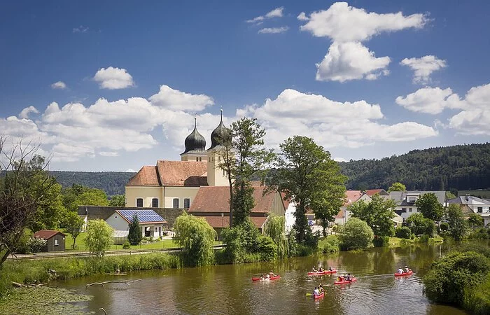 Auf der Altmühl paddeln fünf rote Kanus an Kottingwörth mit ihrer Kirche und deren zwei Kirchtürmen vorbei.
