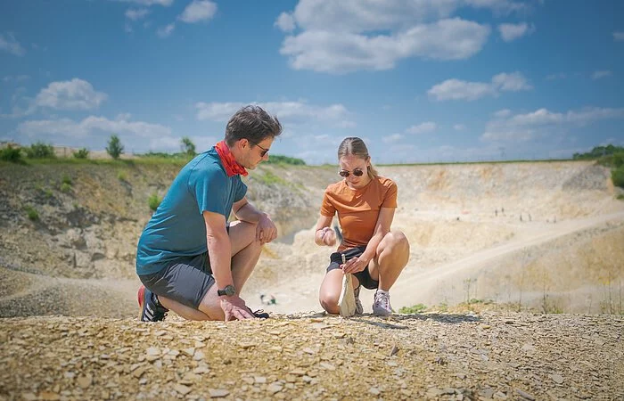 Ein Mann im blauen Shirt kniet neben einer Frau, die mit Hammer und Meißel vorsichtig eine Steinplatte im Fossiliensteinbruch Blumenberg teilt.