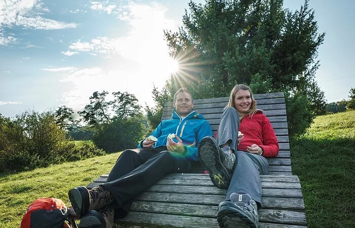 Ein Wanderpärchen sitzt auf einer Liegebank aus Holz. Der Mann mit blauer Jacke hält die Brotzeit in der Hand. Neben ihm sitzt eine blonde Frau mit roter Jacke. Hinter ihnen ist ein großer Busch und die Sonne scheint beiden in den Rücken.