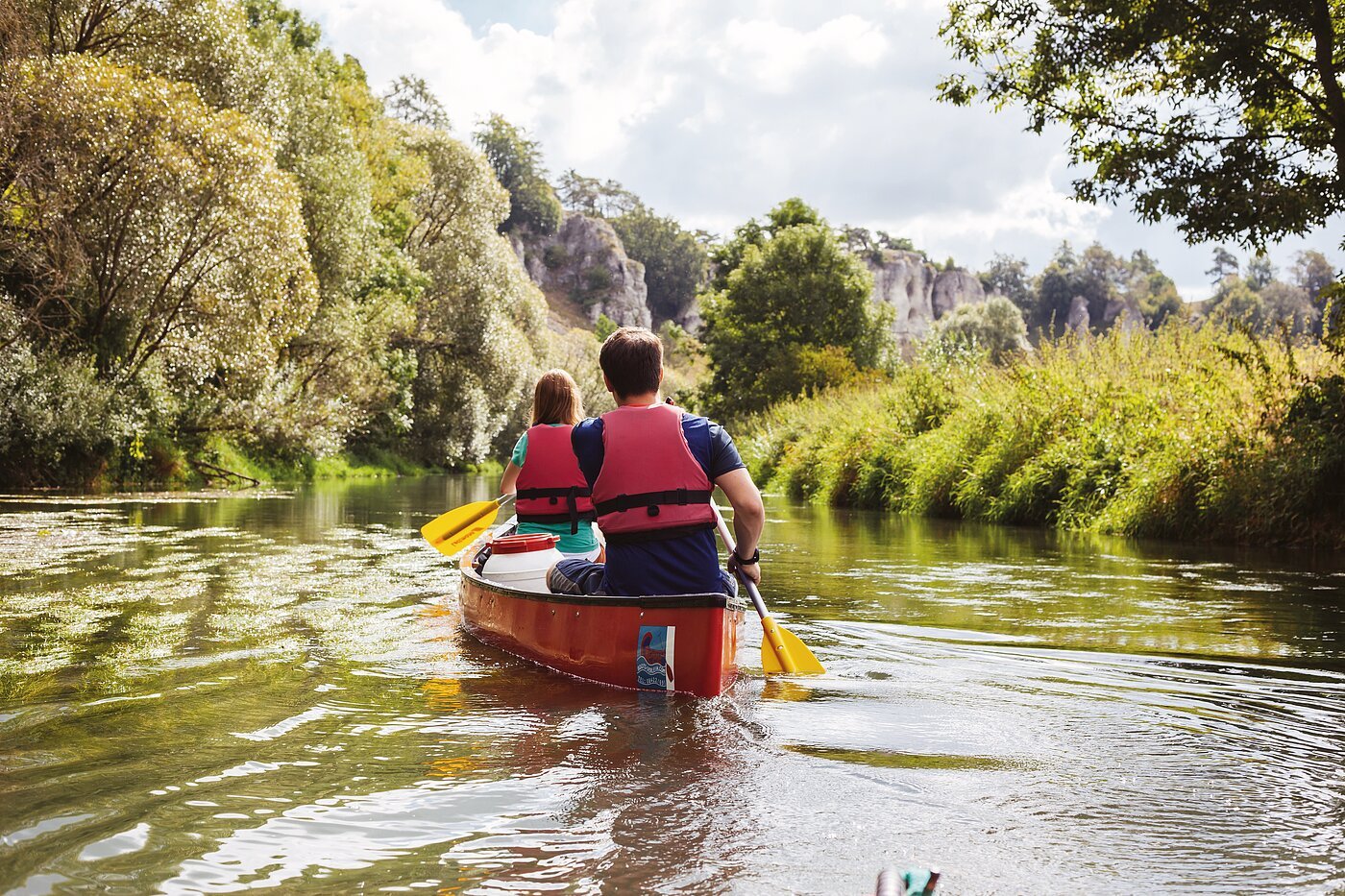 Ein Mann und eine Frau mit dem Rücken zu uns, sitzen in einem roten Kanu auf der Altmühl. Die gelben Paddel erzeugen leichte Wellen auf der Wasseroberfläche. im Hintergrund ist das grüne Ufer mit prächtigen Bäumen zu sehen und rechts blitzt der blaue Himmel durch.