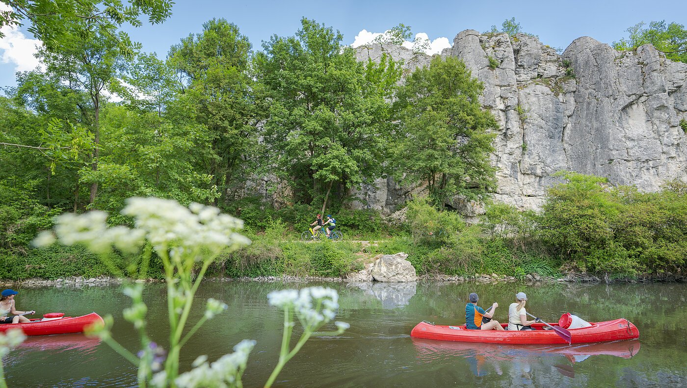 Altmühltal-Radweg bei Dollnstein Zwei rote Kanus auf einem Fluss, im Hintergrund Felsen und Bäume, im Vordergrund unscharfe Blumen.