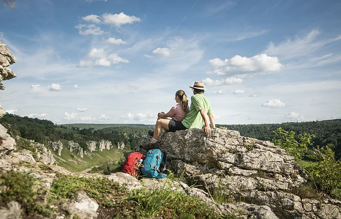 Zwei Wanderer sitzen auf einem Felsen und blicken auf bewaldete Hügel unter blauem Himmel mit Wolken.