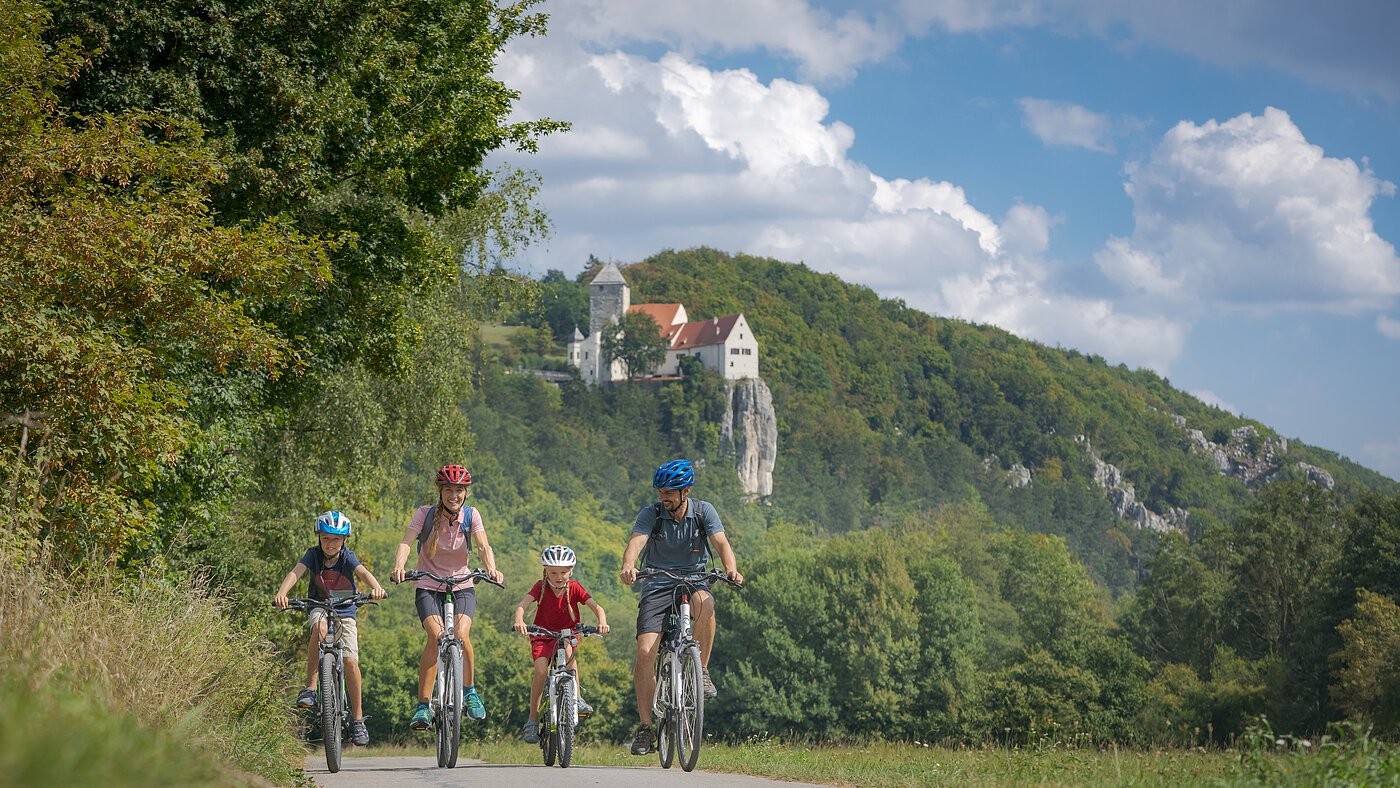 Altmühltal-Radweg (Prunn) Familie mit zwei Kindern fährt auf Fahrrädern auf einem Weg vor bewaldetem Hügel mit Burg im Hintergrund.