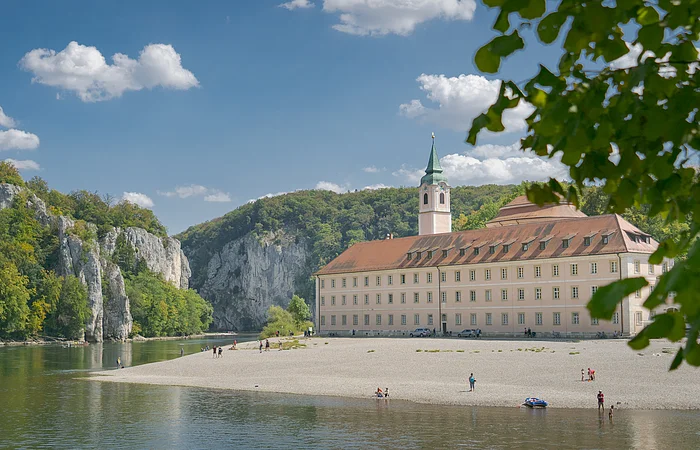 Flussufer mit Kiesstrand, altes Gebäude mit Turm und bewaldete Felsen unter blauem Himmel mit Wolken