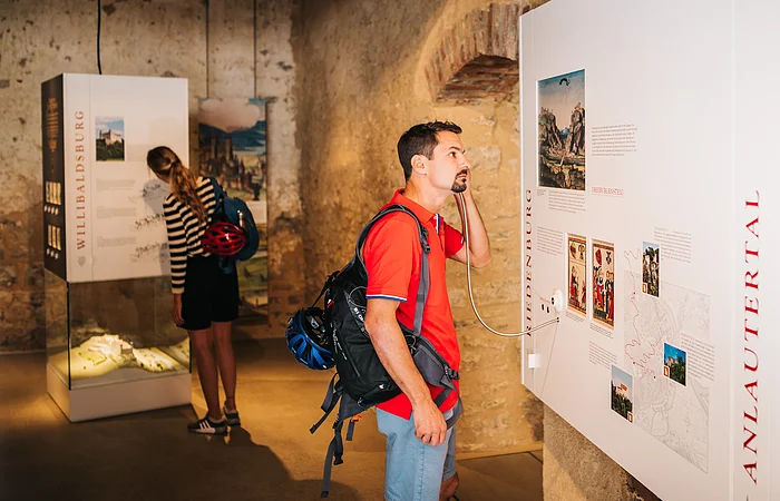 Mann mit rotem Hemd hört an Museumstafel mit Audioguide, Frau liest Ausstellungstext im Hintergrund.