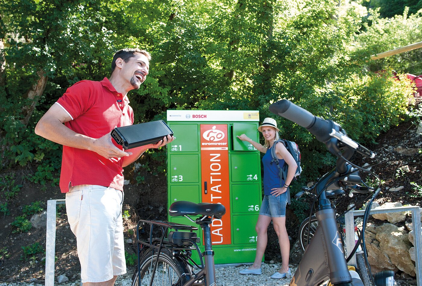 Pärchen am Ladeschrank Mann hält Fahrradakku, Frau öffnet grünen Fahrrad-Ladestation-Schrank im Freien bei Sonnenschein