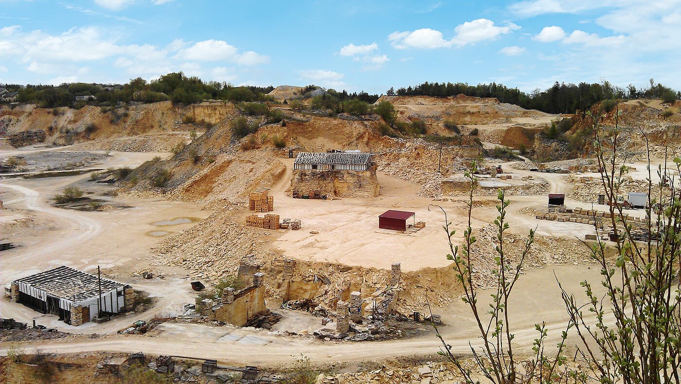 Steinbruch mit Sand- und Felsformationen, Gebäuderuinen und Paletten, umgeben von Vegetation.