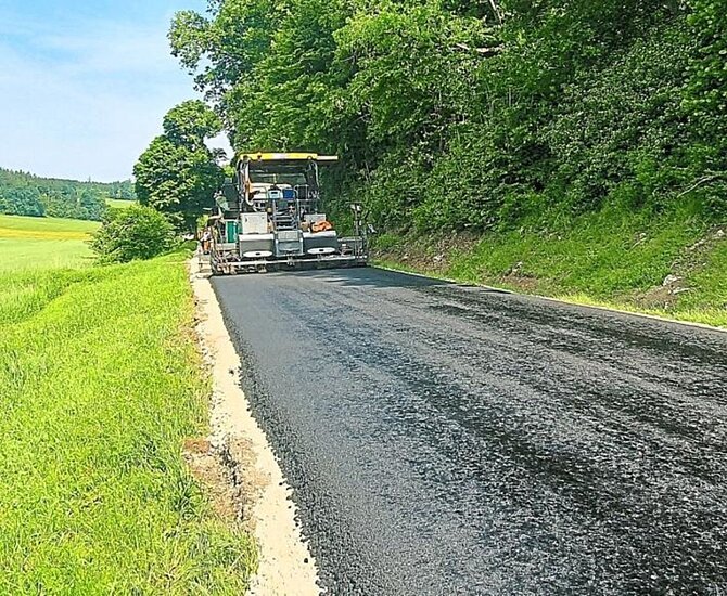 Straße wird mit Baumaschine in ländlicher Gegend neben grünen Wiesen und Bäumen asphaltiert.