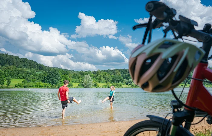 Im Vordergrund stehen rechts zwei Fahrräder, dahinter liegt ein kurzer Strand an einem Seeufer. Im seichten Wasser stehen ein Mann und in eine Frau in kurzen Hosen und T-Shirts, die versuchen, sich gegenseitig nass zu spritzen. Am Ufer gegenüber liegen eine Wiese und Wald.