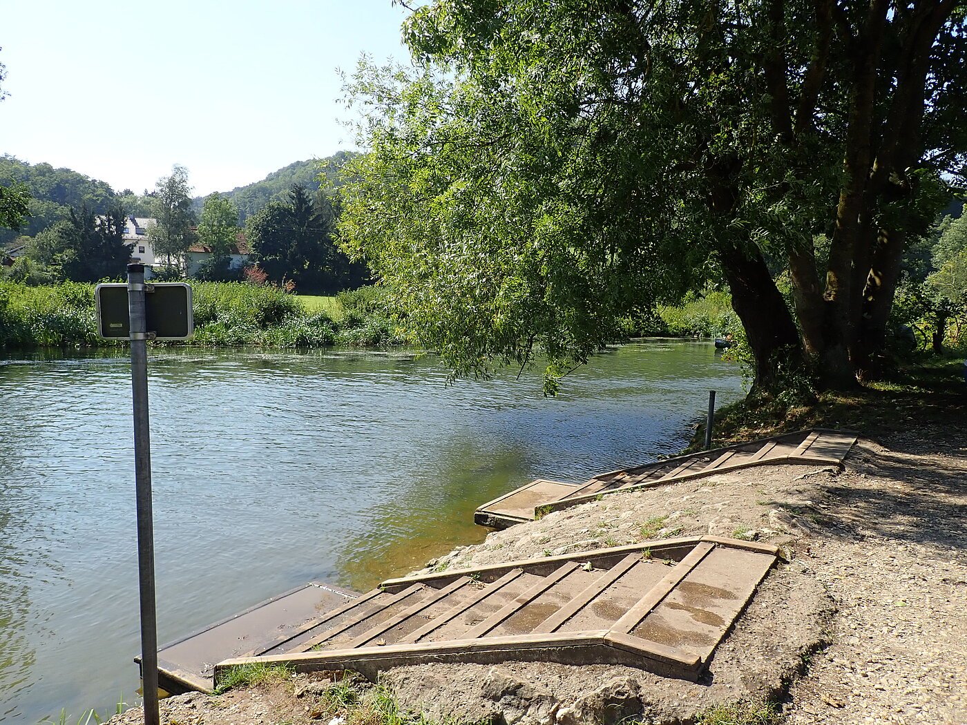 Anlegestelle Hammermühle Ufer mit Treppenstufen zum Wasser, großer Baum rechts, Fluss und Häuser im Hintergrund bei Sonnenschein.